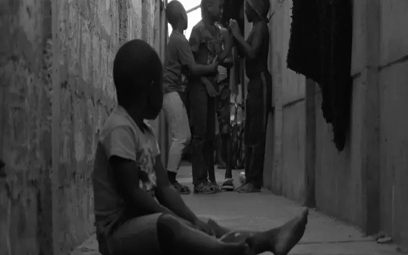 Child sitting in an alley with other children playing in the background.